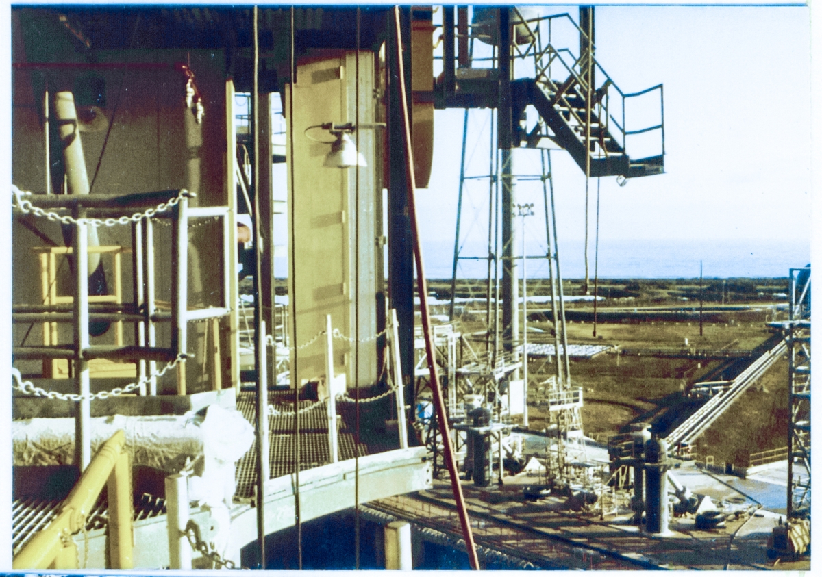 Looking northeast toward the distant Atlantic Ocean from the lowest level of the Space Shuttle Rotating Service Structure at Kennedy Space Center, Florida. In this view the hinged stair that provided access to the orbiter’s left aft fuselage access door can be seen silhouetted against the sky. Behind the stair in the distance, the Sound Suppression Water Tower can be seen. Below, cross-country cryo piping for the LH2 dewar (mostly out of view right of frame), the pad deck, and some of the Flame Trench can be seen, and to the left, platforms and removable handrails of the 112’ elevation of the RSS are visible.